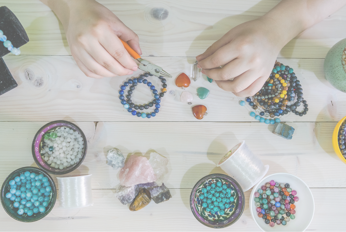 Top-down view of hands crafting beaded jewelry on a light wood table, surrounded by bowls of colorful beads, crystal stones, and spools of thread.